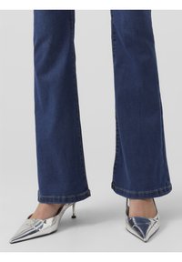 Zapatos de tacón puntiagudos plateados con un acabado elegante y brillante. Llevados con jeans acampanados de color azul oscuro, que revelan el delgado tacón de aguja del zapato.