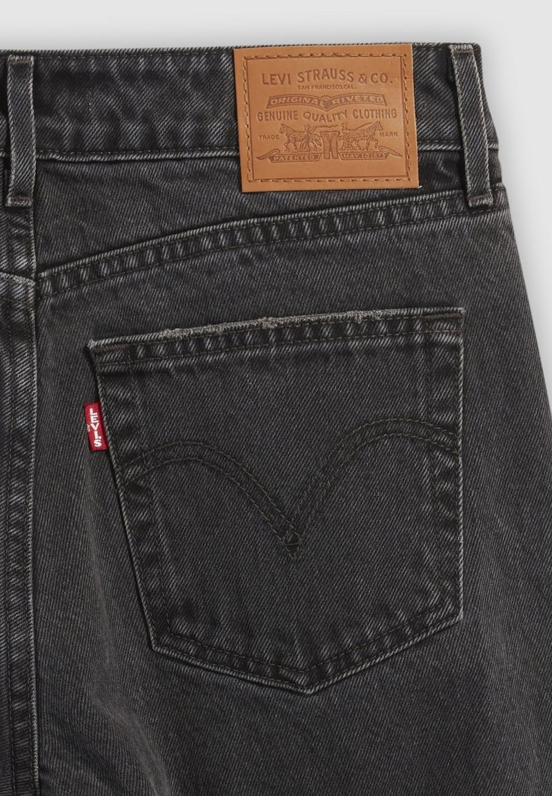 Vaqueros de mezclilla negra con un parche de marca de cuero, costuras visibles y una etiqueta roja característica en el bolsillo trasero. Superficie de tela texturizada.