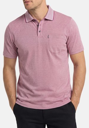 Mann trägt ein kurzärmeliges pinkfarbenes Poloshirt mit Brusttasche und schwarze Hose, Hände in den Taschen, leicht nach links gewandt.