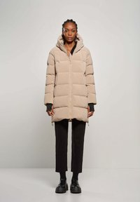 Cappotto imbottito beige con cappuccio, dotato di zip frontale e polsini a coste. Indossato sopra pantaloni neri e scarpe nere chunky.