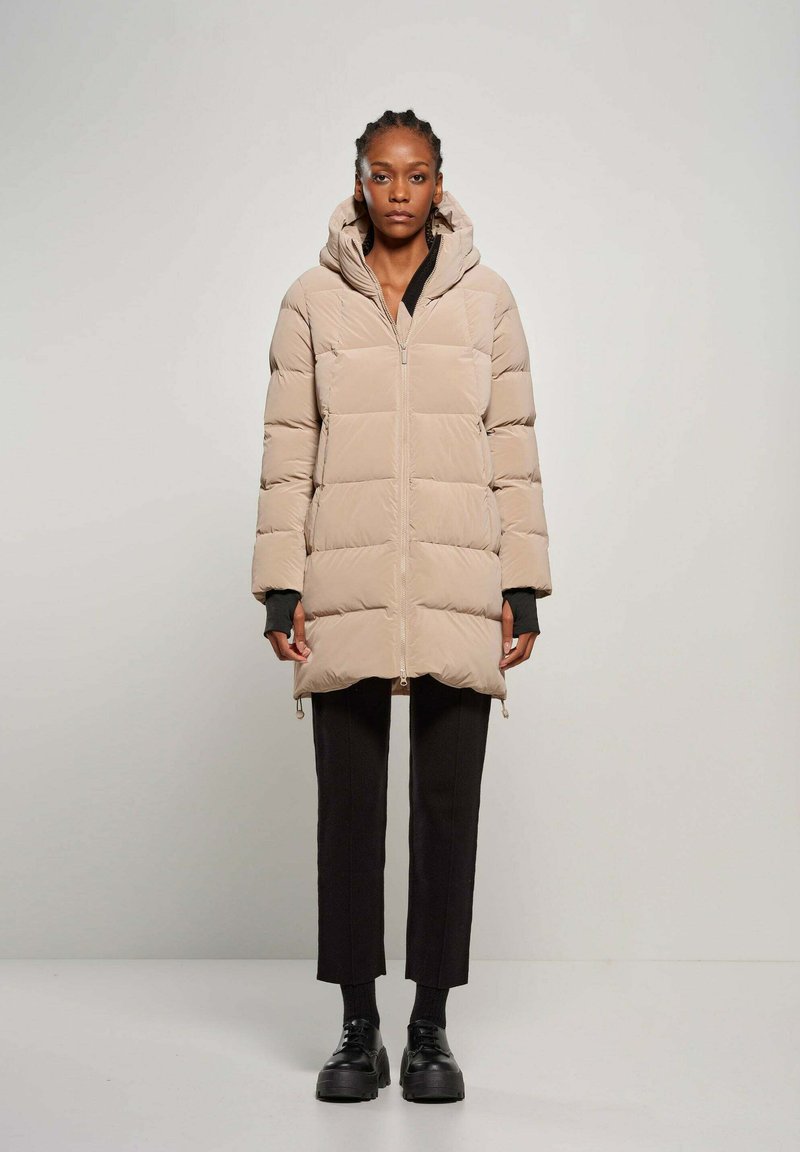 Cappotto imbottito beige con cappuccio, dotato di zip frontale e polsini a coste. Indossato sopra pantaloni neri e scarpe nere chunky.