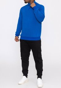 Pull en tricot bleu avec un col montant et des poignets côtelés, associé à un pantalon cargo noir comportant plusieurs poches et des baskets blanches.