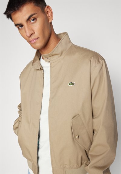 Veste bomber beige en mélange de coton, avec col montant, fermeture éclair sur le devant, poches latérales et logo brodé vert.