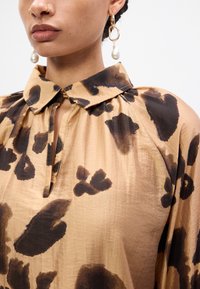 Beige blouse met een luipaardprint in donkerbruin. Beschikt over een staande kraag en een sleutelgatoopening bij de hals. Soepele textuur.