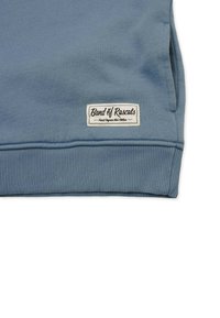 Sininen huppari, jossa on ribbireunat ja teksturoitu pinta. Alaosaan on ommeltu valkoinen etiketti, jossa on musta teksti.