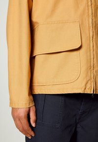 Tan jacket with a zip front and large flap pocket. Fabric appears smooth with a subtle texture. Worn over dark pants.