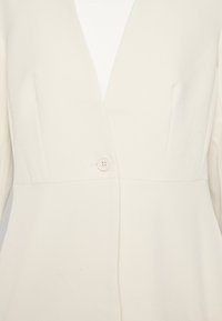 Blazer couleur crème avec une coupe ajustée, présentant un décolleté en V profond, une fermeture à un bouton, et un tissu lisse légèrement texturé.