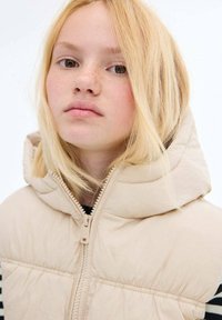 Beige Steppweste mit hohem Kragen und Kapuze, ausgestattet mit einem Reißverschluss auf der Vorderseite und gepolsterten Texturen; über einem gestreiften Hemd getragen.