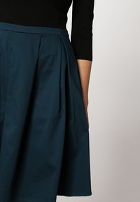 Una falda pliseada en color verde azulado, hecha de una tela de textura suave, combinada con una blusa negra ajustada con mangas de tres cuartos.