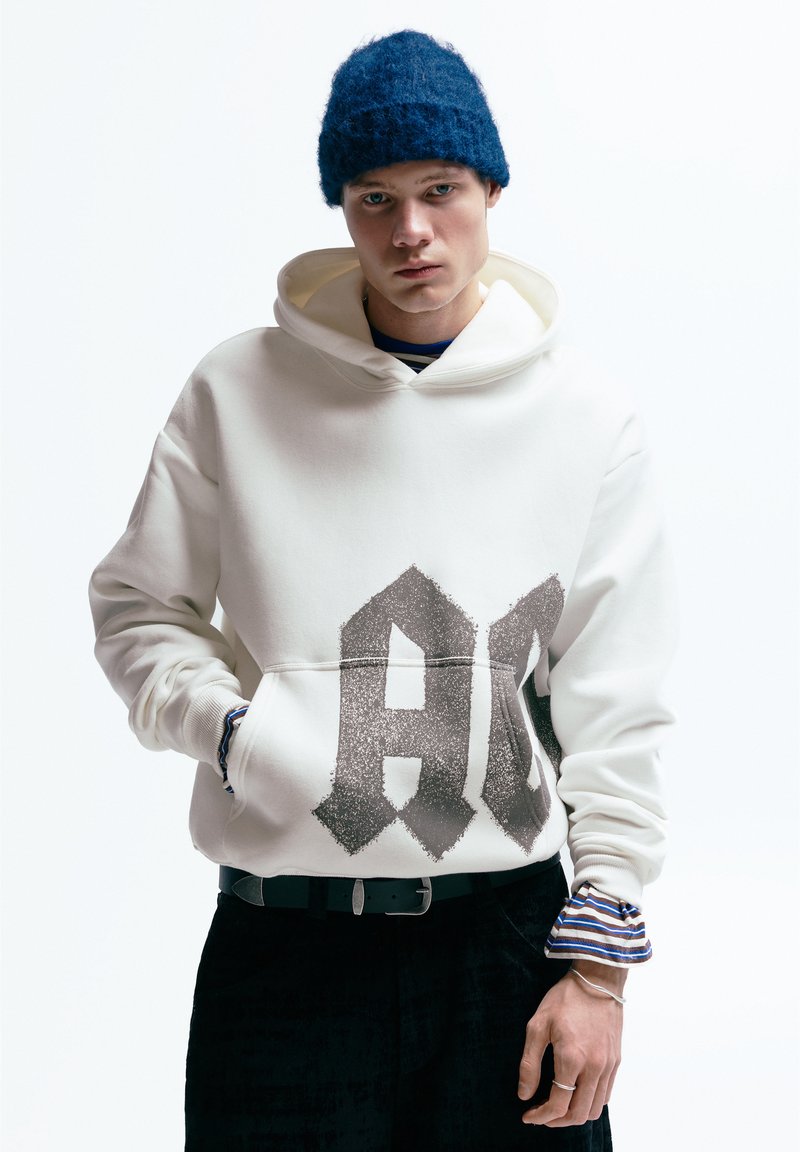 Sweat à capuche blanc avec un grand logo imprimé en gris, équipé d'une poche kangourou. Un pantalon noir et un bonnet bleu texturé complètent la tenue.