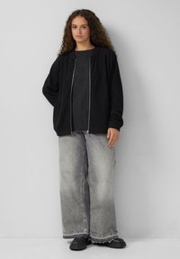 Vrouw met krullend haar, gekleed in een zwarte ritsjack, een donkere trui, brede grijze jeans en zwarte chunky laarzen, staande tegen een egale muur.