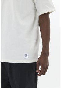 Weißes kurzärmeliges T-Shirt mit lockerer Passform, versehen mit einem kleinen etikettierten Etikett mit der Aufschrift „M 73“ am Saum, kombiniert mit dunkler Hose.