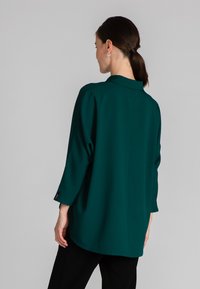 Blusa verde, con texture, dal taglio morbido, colletto classico e maniche tre quarti. Tessuto liscio e orlo leggermente curvo.