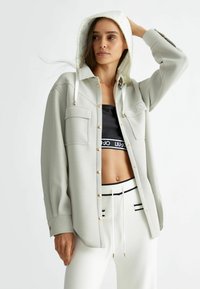 Heledhallane hallikas jope kuldsete nööpidega, millel on kaks rinna taskut. Üks all, must crop-top logoga ja triibulised valged spordipüksid.