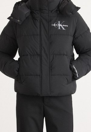 Doudoune noire matelassée avec poches avant, logos Calvin Klein Jeans sur la poitrine et la manche, portée avec un pantalon noir.