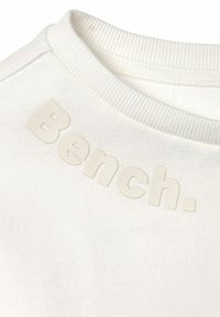 Felpa bianca con un colletto a costine, caratterizzata da un logo testurizzato leggermente in rilievo che recita "Bench." sul lato sinistro superiore.