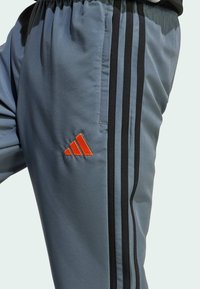Grå träningsoverall med svarta sidosträck och en orange Adidas-logotyp. Tillverkad av mjukt tyg, med elastisk midja och fickor.