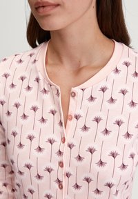 Vrouw in een lichtroze knoopshirt met een donkerroze abstracte bloemenpatroon en een ronde kraag, waarbij haar nek en een deel van haar gezicht zichtbaar zijn.