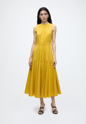 Femme debout portant une robe midi jaune sans manches avec une taille froncée et des sandales à talons noires sur fond blanc uni.