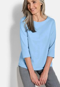 Helles blaues, Rundhalsoberteil mit Dreiviertelärmeln, aus weichem Stoff, kombiniert mit grauen Hosen, vor einem neutralen Hintergrund.