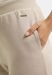 Beżowe dresy z ribbowanym paskiem w talii, z boczną kieszenią i małym metalowym logo. Miękka tekstura z gładkim wykończeniem.