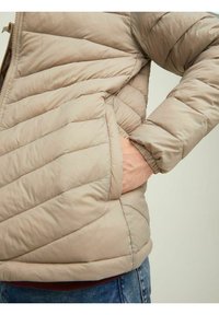 Beige padaná bunda s horizontálním prošíváním, zipovým zapínáním a bočními kapsami. Manžety a lem jsou žebrované, vyrobená z lehkého syntetického materiálu.