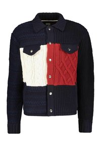 Stickad jacka i marinblått, rött och vitt blockmönster. Har en krage, knäppning fram och två bröstfickor med silverknappar. Texturerade mönster.