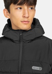 Giacca nera imbottita con colletto alto e chiusura con zip. Presenta un logo ricamato sul petto e una texture in tessuto liscia e isolante.