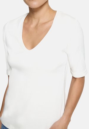 Femme portant un t-shirt blanc uni à col en V avec des manches jusqu'au coude, debout avec un bras légèrement plié.