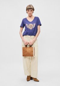 Μπλε t-shirt με v-neck και πολύχρωμο σχέδιο, μπεζ παντελόνι με φαρδιές γραμμές και μια μικρή καφέ τσάντα tote με υφή στα πλάγια και κοντές λαβές.