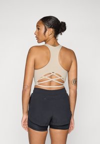 Beige sportsbh med krysset design på baksiden, brukt med svarte shorts. Har en glatt tekstur og tettsittende silhuett.