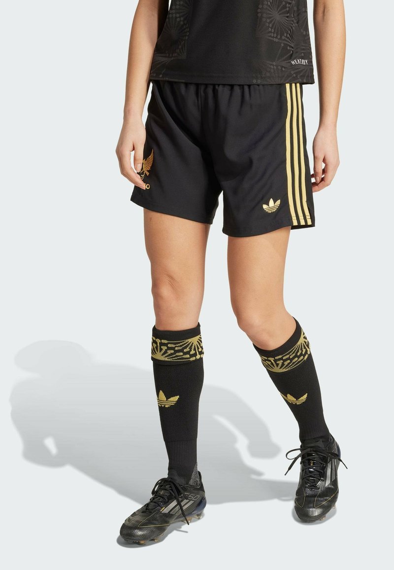 Pantaloni sportivi neri con strisce laterali dorate e logo; abbinati a calze nere fino al ginocchio con dettagli dorati e un scarpetta da calcio nera.