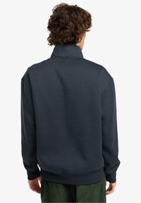 Granatowy sweter z wysokim kołnierzem, długimi rękawami oraz ściągaczami na mankietach i u dołu. Gładka tekstura, luźny krój i minimalne zdobienia.