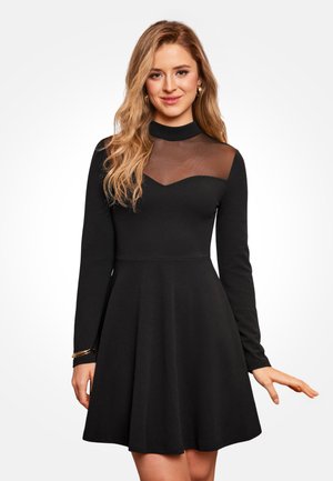 Robe noire à manches longues avec un corsage ajusté, un panneau en mesh transparent au niveau de l'encolure, et une jupe évasée, avec un subtil accent doré sur la manche.
