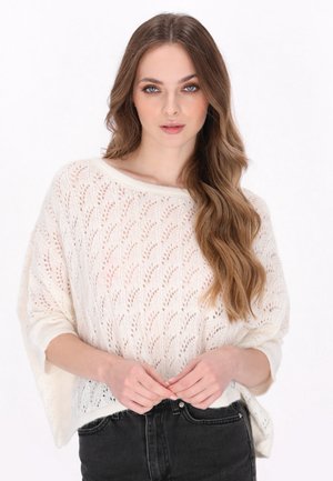 Hvid strikket sweater med et blomstermønster, afslappet pasform og bred bærestrop. Båret over sorte højtaljedebukser.