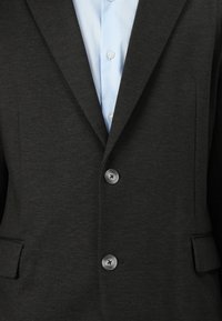 Dark gray blazer with a textured finish, featuring a notched lapel, two buttons, and side pockets; paired with a light blue shirt.