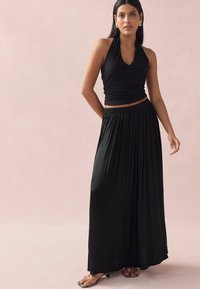 Crni halterneck crop top s V-izrezom, uparen s lepršavom crnom maksi suknjom s pliseima. Model koji nosi ravne sandale stoji.