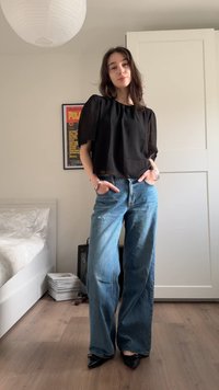 Femme debout dans une pièce, portant un chemisier noir, un jean large bleu, et des chaussures pointues noires, les mains dans les poches.