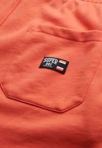 Tessuto color corallo con una texture morbida, caratterizzato da un'etichetta nera con la scritta "SUPER DRY ATHLETICS" e strisce arancioni e bianche. Dettaglio della tasca.