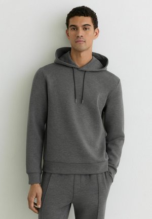 Joven con el pelo rizado y corto que lleva una sudadera con capucha gris oscuro y pantalones deportivos a juego, de pie frente a una pared lisa y clara.