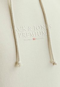 Sudadera beige de tela texturizada, con un logo elevado en blanco que dice "JACK & JONES PREMIUM" y cintas ajustables con extremos anudados.