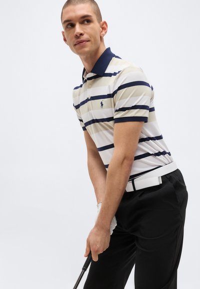 Jeune homme en polo rayé et pantalon noir tenant un club de golf, prêt à faire un swing sur un fond uni.