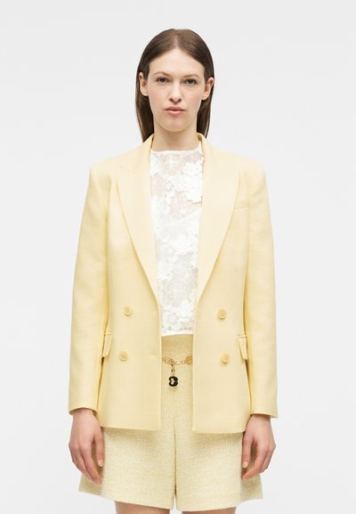 Femme portant un blazer croisé jaune pâle, un haut en dentelle florale blanche transparente, et un short texturé jaune assorti avec une ceinture chaîne.