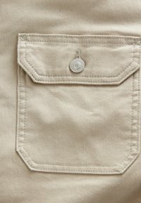Beige stoflomme med rektangulær form, udstyret med en sølvfarvet trykknap og kontrasterende syninger.
