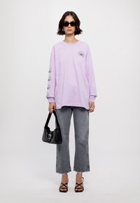 Camisa de manga larga lavanda con estampado de logo, combinada con jeans acampanados gris desgastados y sandalias negras con tiras. Un bolso negro completa el look.