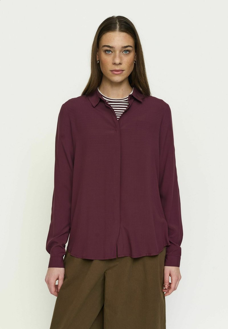Femme aux cheveux bruns et lisses portant un chemisier violet à manches longues sur une chemise rayée et un pantalon marron à jambes larges, debout devant un fond uni.