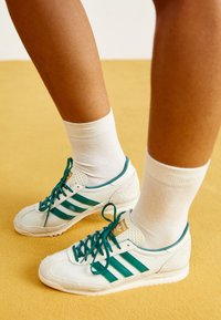 Vita sneakers med gröna ränder, snörning, gummisula, detaljer i mocka och matchade med vita crew-sockor mot en gul bakgrund.