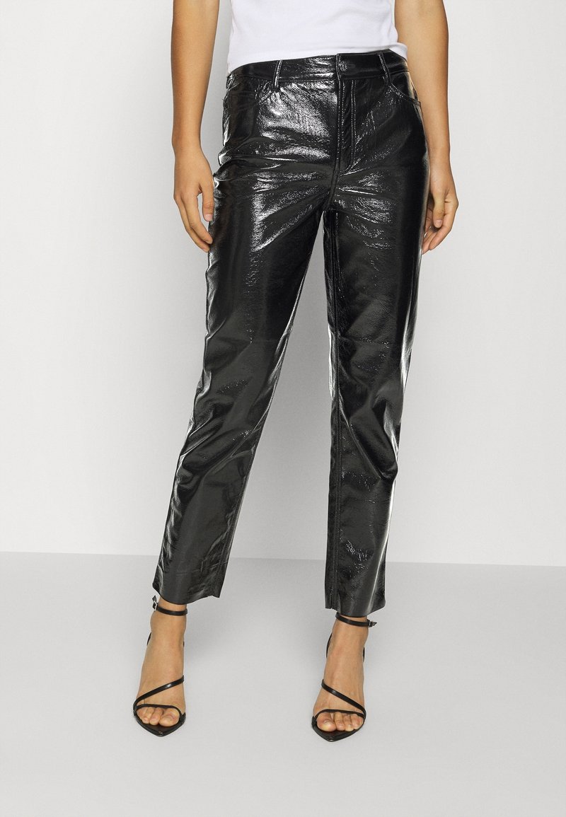Personne portant un pantalon en cuir noir brillant à jambes droites et des sandales noires à talons hauts avec des brides, debout sur un fond uni.