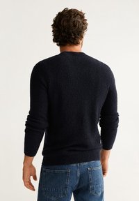 Uomo con capelli ricci che indossa un maglione scuro e testurizzato e jeans blu, girato di spalle su uno sfondo chiaro e uniforme.