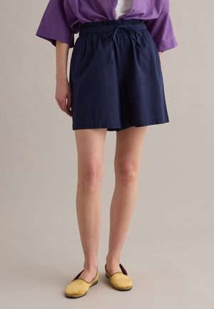 Persoon die marineblauwe shorts met trekkoord, gele instapschoenen en een paars shirt met opgerolde mouwen draagt, staand op een effen achtergrond.